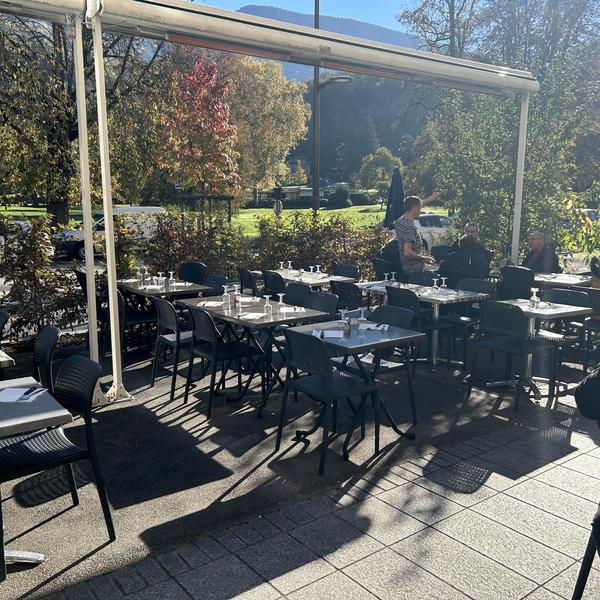 Terrasse de café/restaurant ensoleillée avec une vue sur un parc avec des grands arbres.